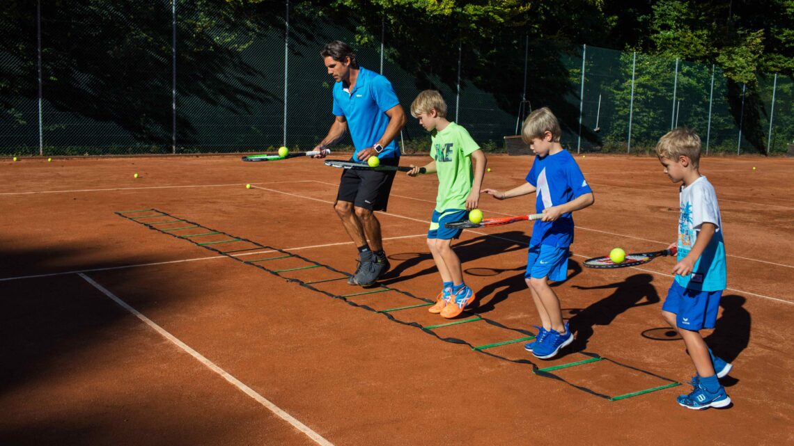 Trainer Sven Melig zeigt Kindern bei Kids Sportcamps auf dem Tennisplatz Geschicklichkeitsübungen mit Schläger und Ball.