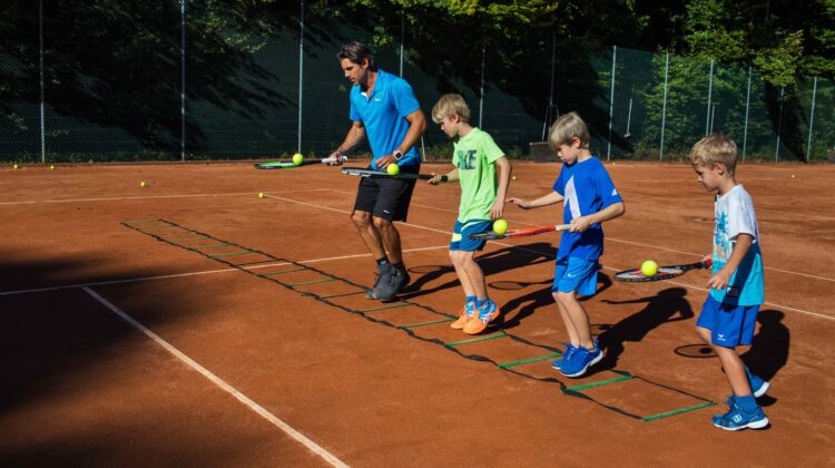 Trainer Sven Melig zeigt Kindern bei Kids Sportcamps auf dem Tennisplatz Geschicklichkeitsübungen mit Schläger und Ball.